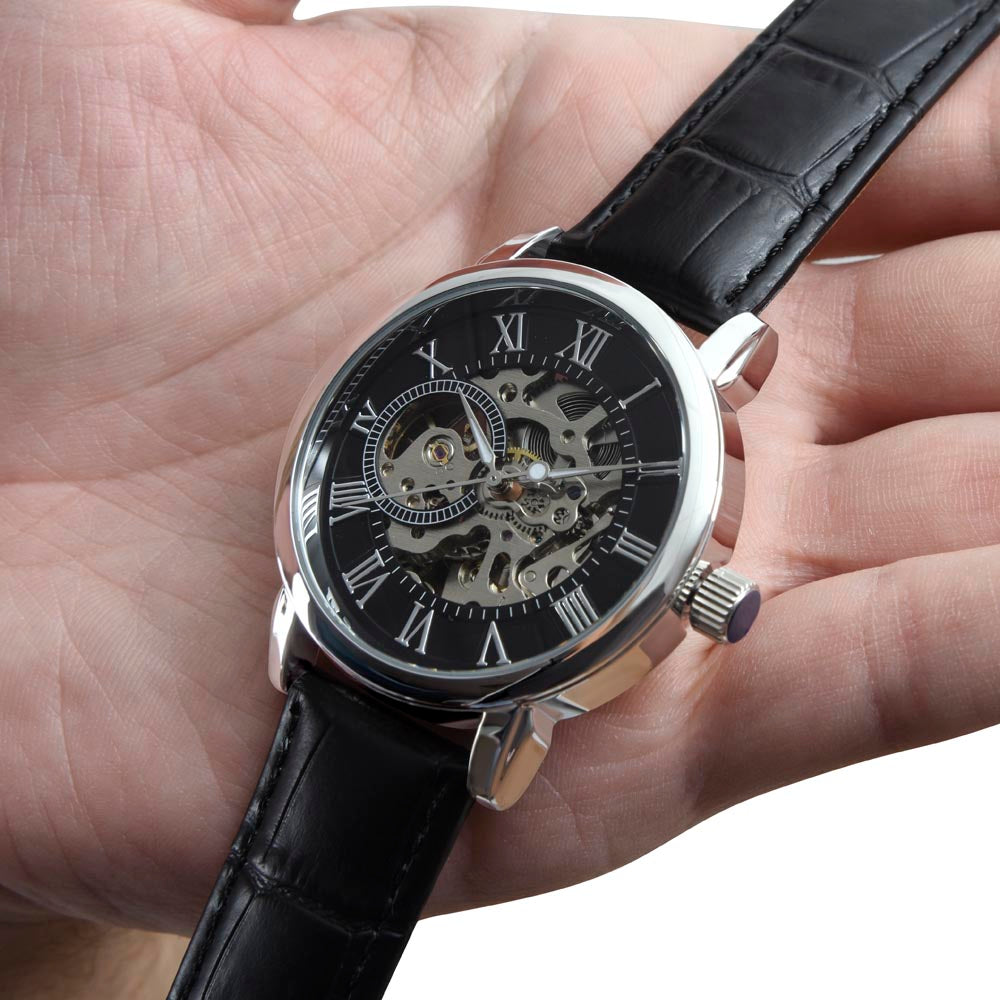 Reloj de pulsera esqueleto para hombre con correa de cuero negro con textura de cocodrilo, sostenido sobre la palma de una mano para mostrar su tamaño real.