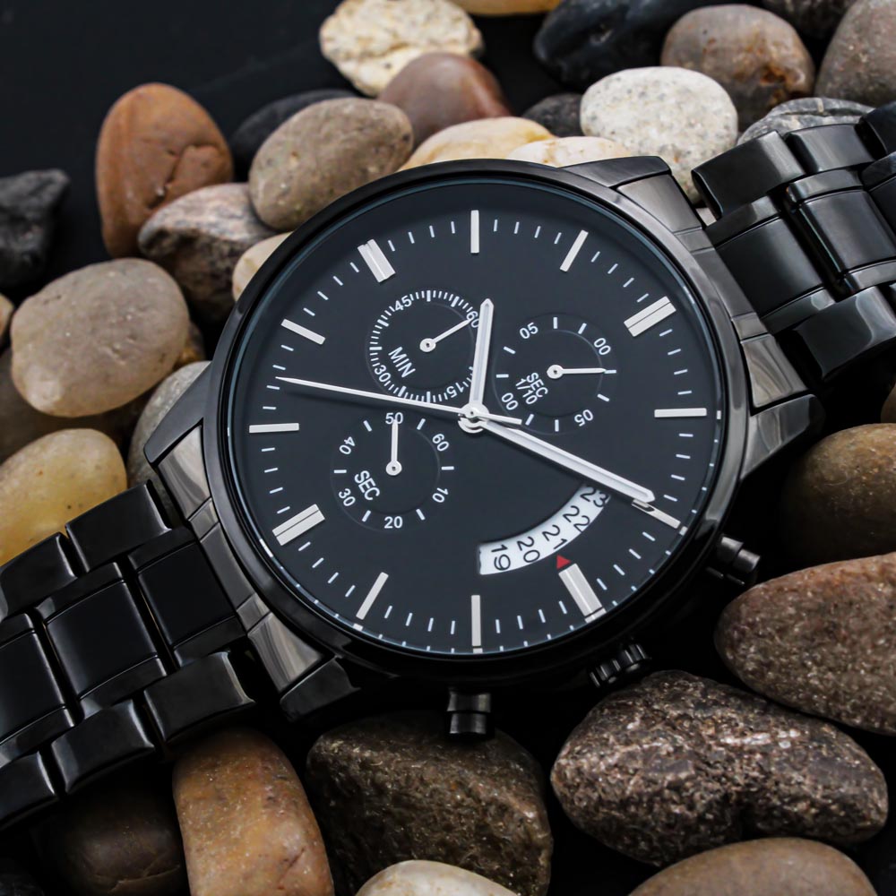 Black stainless steel men's chronograph watch with date and analog time with a white dial laying on a bed of various rocks.