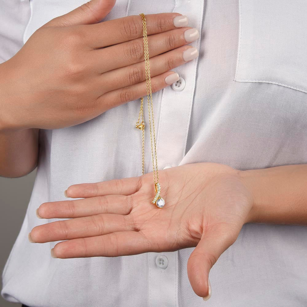 A woman wearing a white button-up shirt holds a gold Alluring Beauty pendant necklace draped between both of her hands. The delicate gold cable chain loops over her upper hand, while the small ribbon-shaped pendant with its round sparkling crystal center rests gently in the palm of her open lower hand.