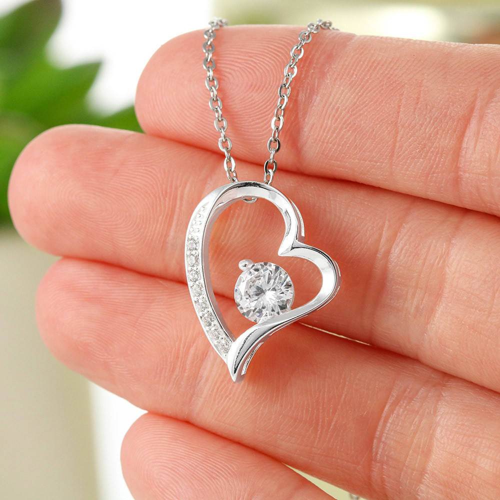Collar con colgante de plata en forma de corazón, con una gema central redonda y brillante y piedras de acento, mostrado sobre los dedos de una persona.