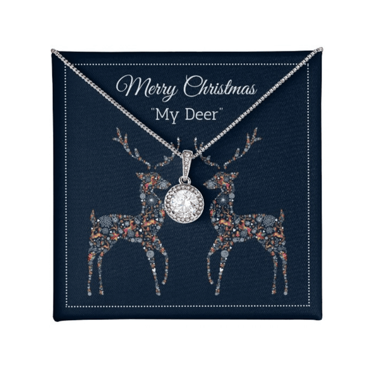 A silver-toned Christmas necklace with a round crystal-studded pendant, displayed on a navy blue gift card that reads "Merry Christmas 'My Deer'" and features two decorated deer illustrations.