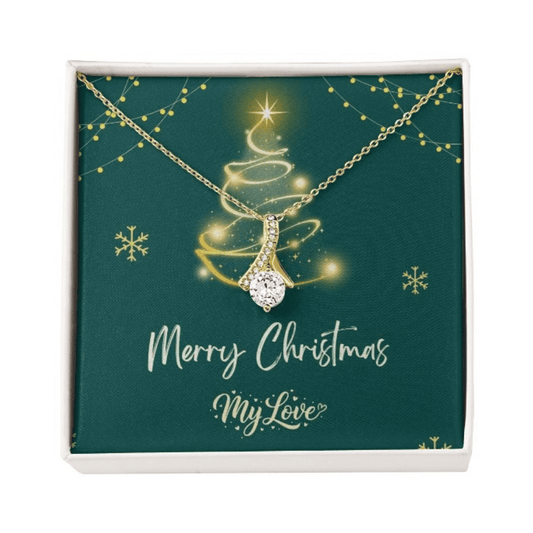 A gold Christmas tree pendant necklace with a sparkling clear teardrop-shaped center stone, presented on a dark green square gift card inside a white box. The card features a gold glittery Christmas tree, snowflakes, and the text 'Merry Christmas My Love