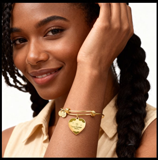 A close-up lifestyle shot of a woman wearing a gold bangle bracelet with a heart-shaped charm engraved with 'Love Always' and a small tag, set against a clean white background.