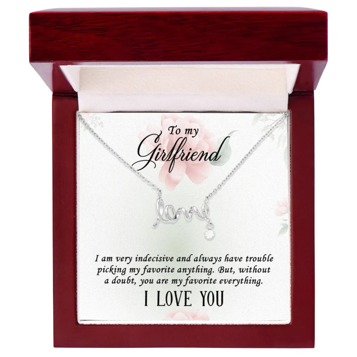 A silver 'I love you' necklace with a dangling 'love' charm is presented in an open dark red wooden gift box. The necklace is displayed on a white card with a romantic message for a girlfriend.