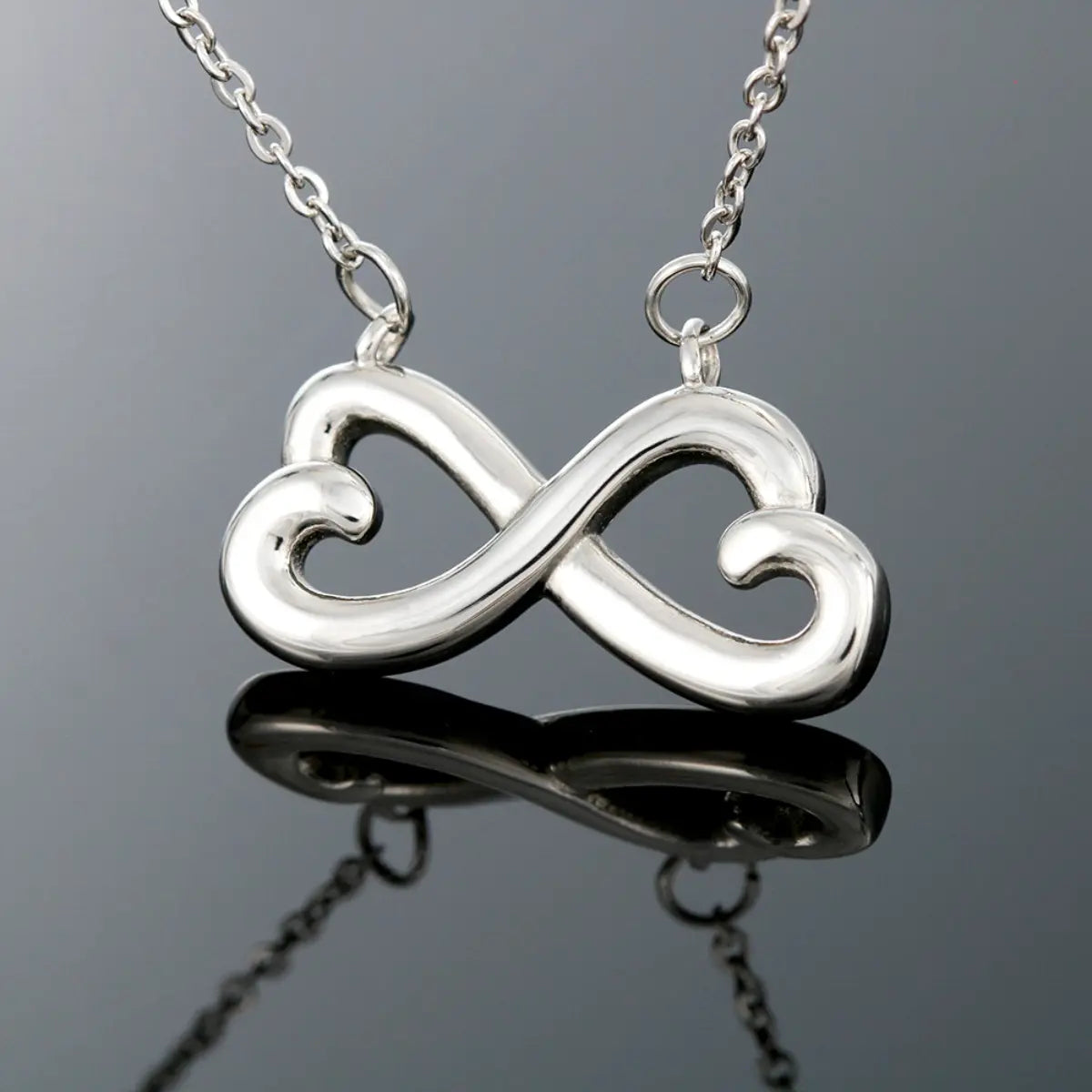 Silver infinity symbol necklace on a reflective surface with a gray background