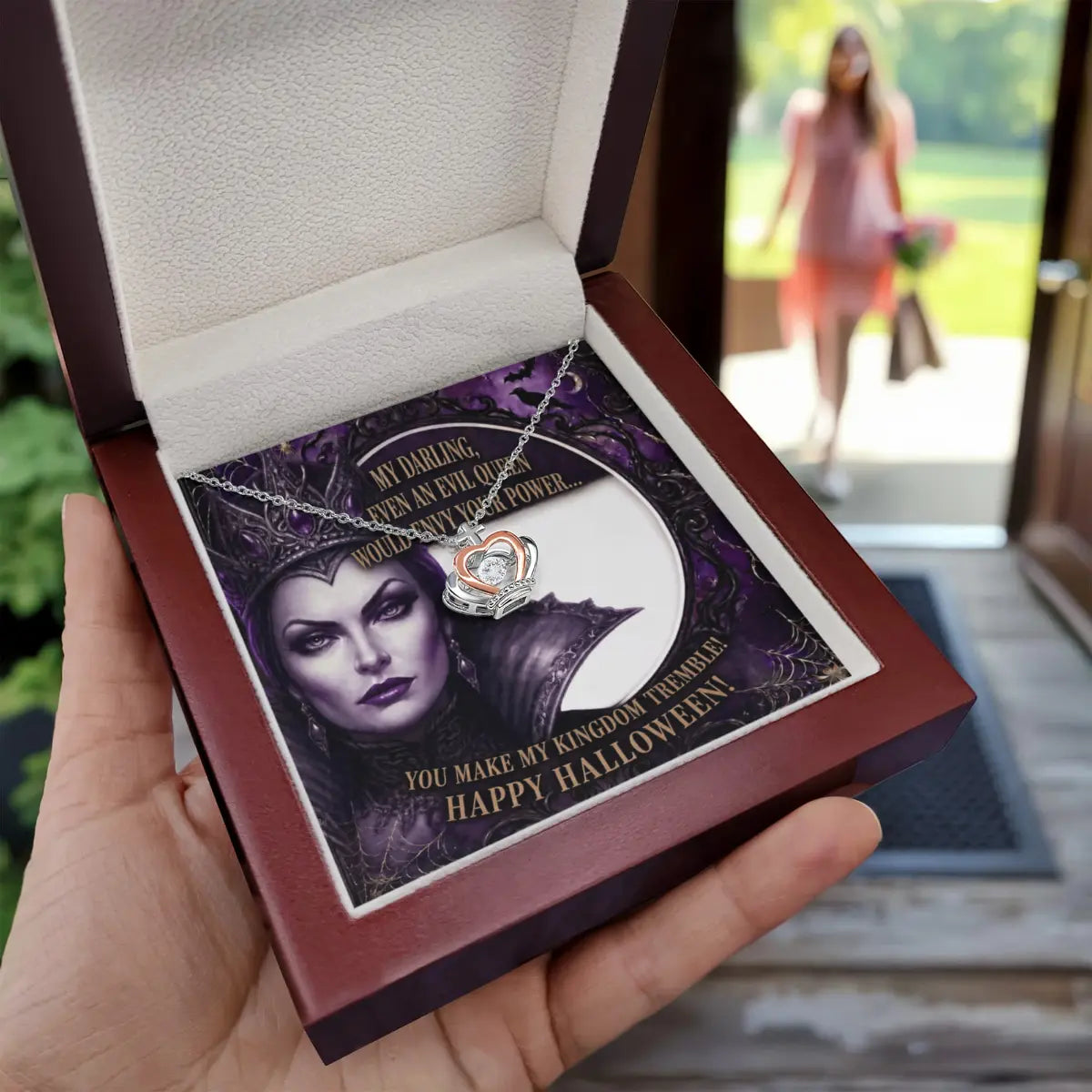 A hand holds an open dark wood gift box containing a necklace with a silver and rose gold crown pendant with cubic zirconia on a printed card. The card features a portrait of Maleficent and reads, 'My Darling, You Are An Evil Queen With Any Power You Want! Happy Halloween!'.