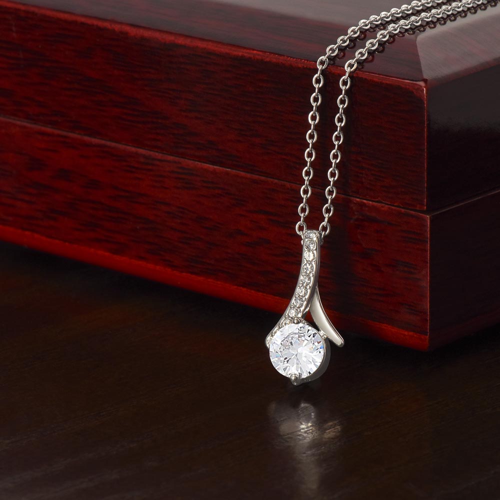 An 14k white gold finish Alluring Beauty pendant necklace rests against a dark red wooden gift box on a dark wooden surface. The pendant features a large, brilliant round-cut cubic zirconia crystal at the bottom, suspended from a curved, ribbon-like bail lined with delicate pavé-set accent crystals. The sparkling pendant is attached to a classic cable chain.