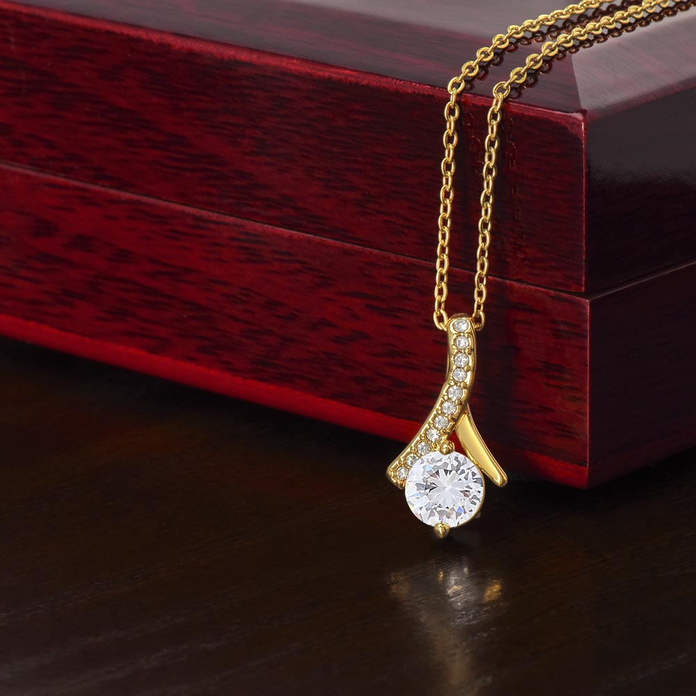 An 18k yellow gold finish Alluring Beauty pendant necklace rests against a dark red wooden gift box on a dark wooden surface. The pendant features a large, brilliant round-cut cubic zirconia crystal at the bottom, suspended from a curved, ribbon-like bail lined with delicate pavé-set accent crystals. The sparkling pendant is attached to a classic cable chain.