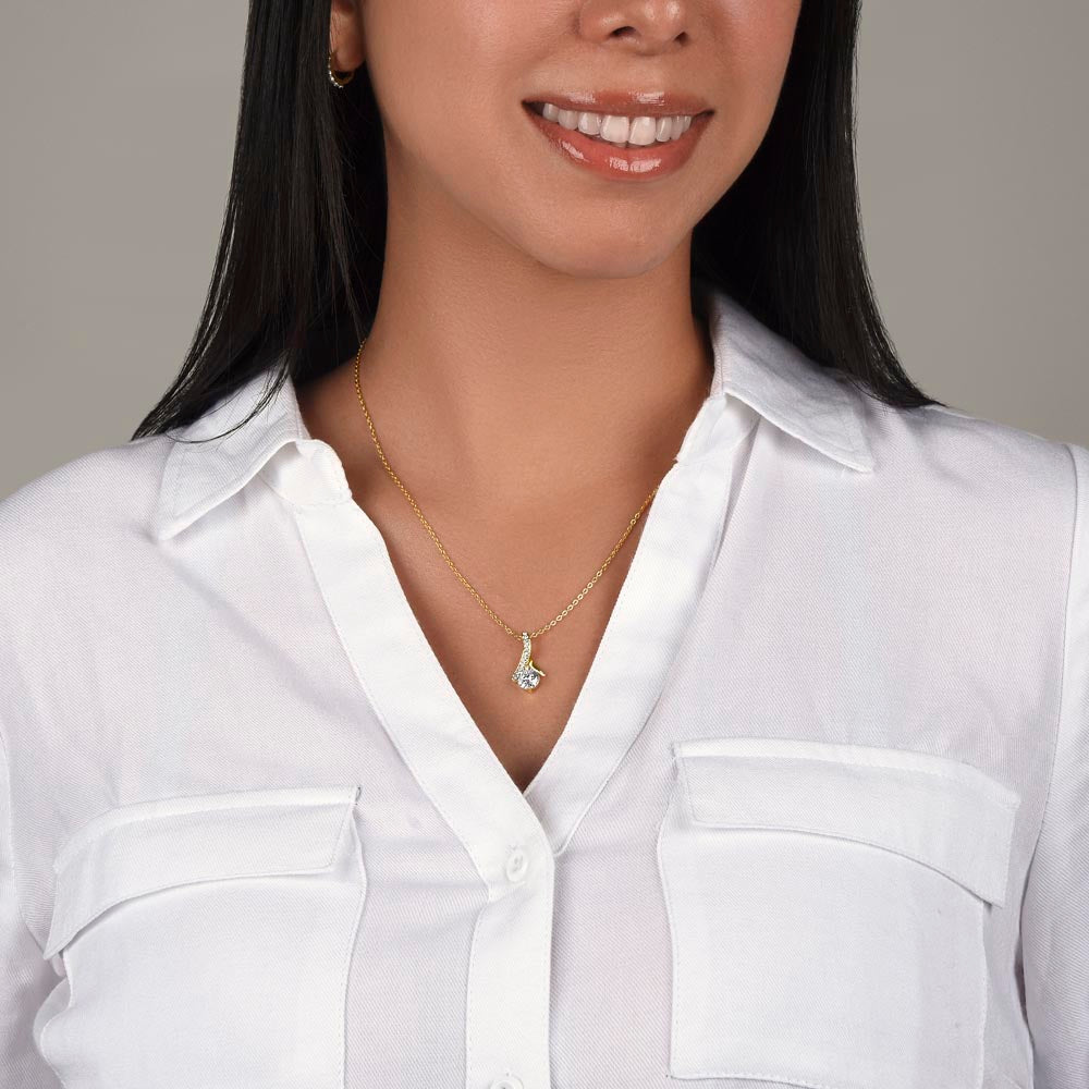 A close-up view of a smiling woman with dark hair wearing a white button-up shirt and modeling a gold Alluring Beauty pendant necklace. The delicate gold cable chain sits elegantly around her collarbone, showcasing the small, ribbon-shaped pendant and its sparkling, round crystal center.