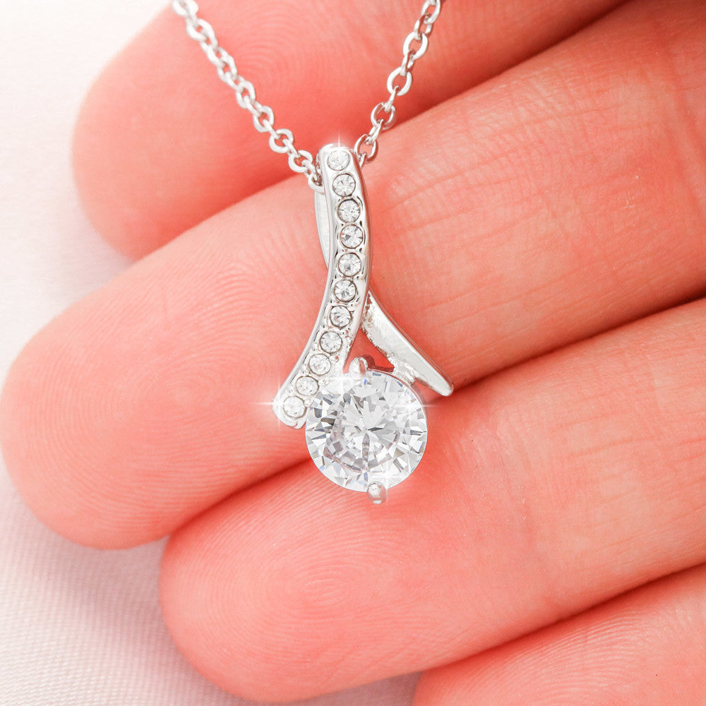 A close-up shot of a silver Alluring Beauty pendant necklace gently resting across a person's fingers. The pendant features a large, brilliant round-cut cubic zirconia crystal at the base, topped with a curved ribbon-style bail lined with a row of sparkling pavé-set accent crystals. The pendant is suspended from a delicate cable chain.