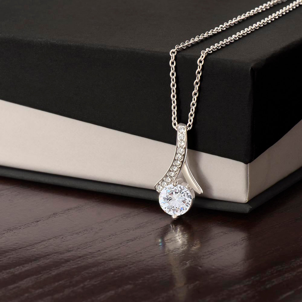 A 14k white gold finish Alluring Beauty pendant necklace rests against a black and white gift box on a dark wooden surface. The pendant features a large, brilliant round-cut cubic zirconia crystal at the bottom, suspended from a curved, ribbon-like bail lined with delicate pavé-set accent crystals. The sparkling pendant is attached to a classic cable chain.