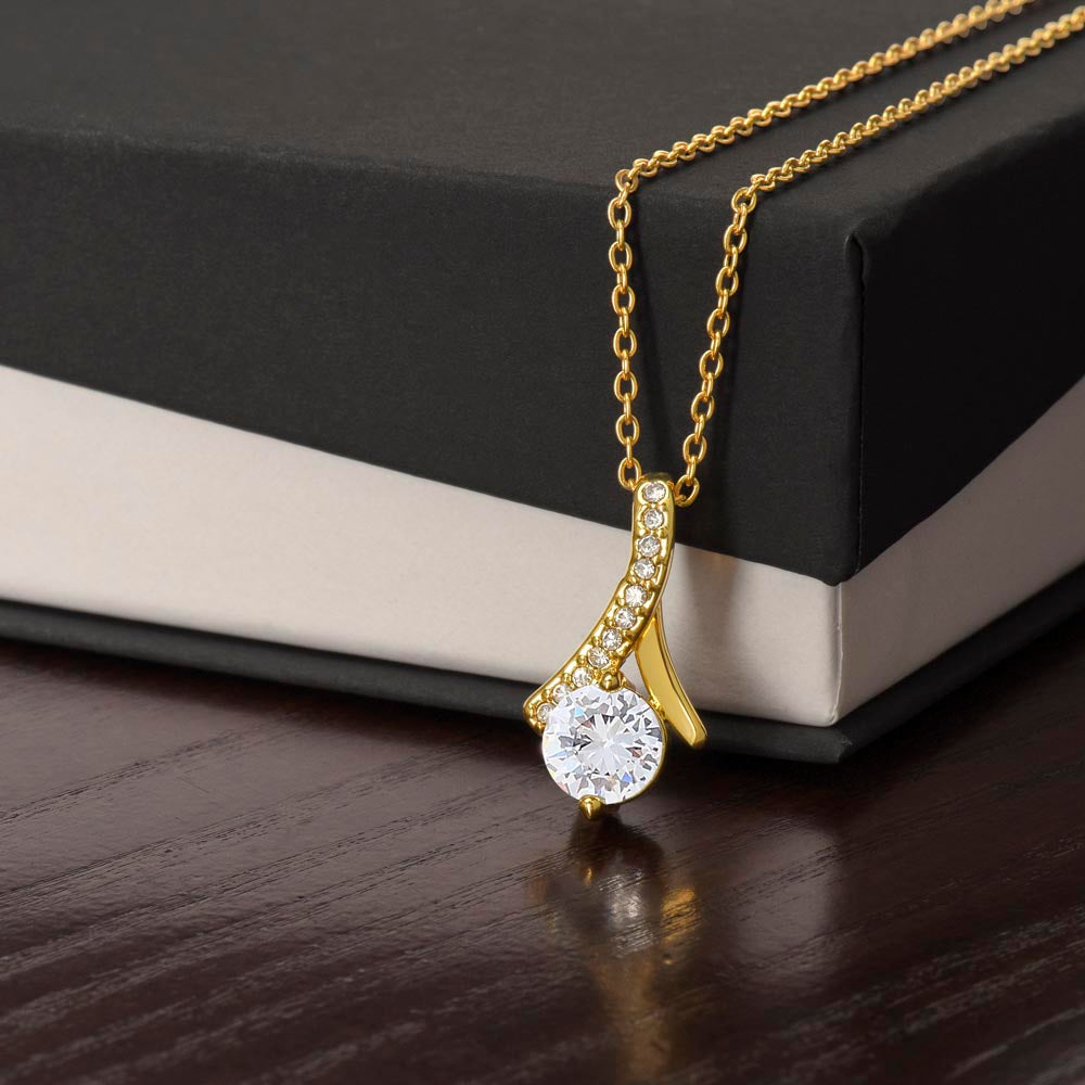 An 18k yellow gold finish Alluring Beauty pendant necklace rests against a black and white gift box on a dark wooden surface. The pendant features a large, brilliant round-cut cubic zirconia crystal at the bottom, suspended from a curved, ribbon-like bail lined with delicate pavé-set accent crystals. The sparkling pendant is attached to a classic cable chain.
