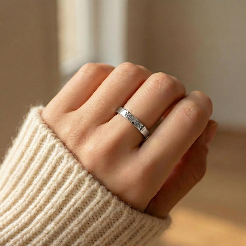 A close-up lifestyle photo of a person wearing a polished silver stainless steel flat-profile band ring on their ring finger. The person's hand is tucked cozy against a soft, cream-colored ribbed knit sweater sleeve. The ring features a crisp, centered sample engraving that reads "Your Text" along the band, set against a softly blurred, warm interior background.