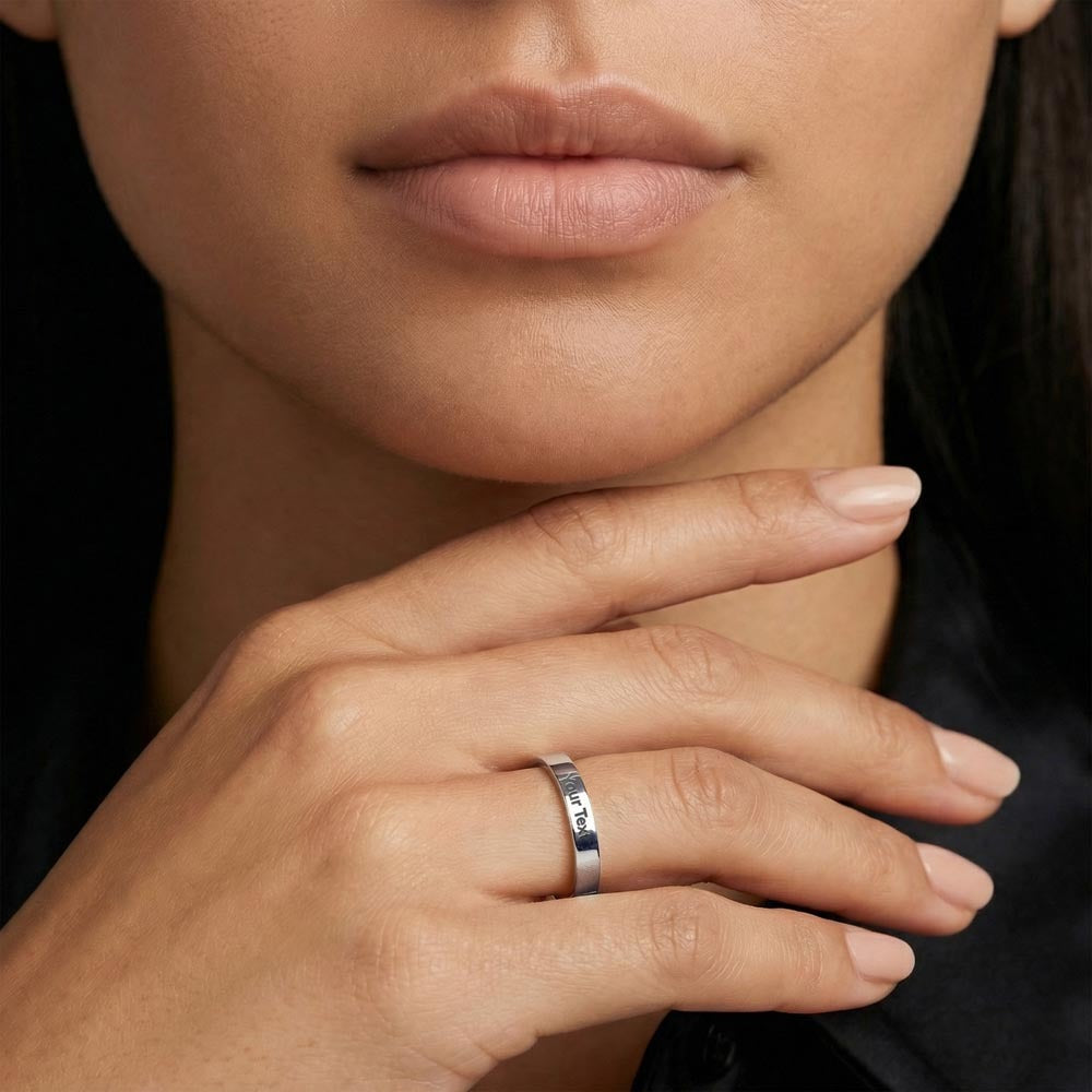 A close-up lifestyle photo of a woman resting her hand gracefully beneath her chin, showcasing a polished silver stainless steel flat-profile band ring on her finger. The customizable ring features a clean sample engraving that reads "Your Text" along the band. She is wearing a black shirt, set against a dark background that highlights the jewelry.