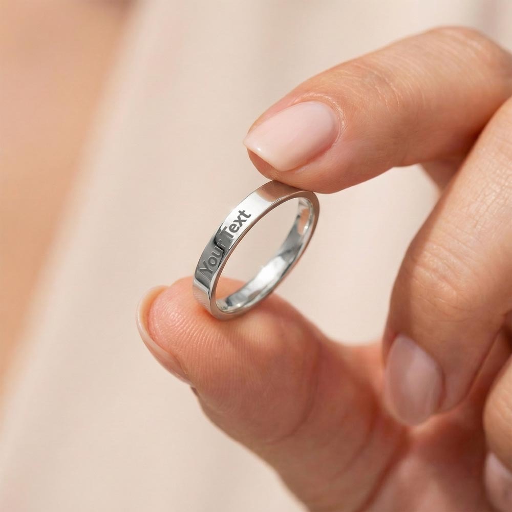 A close-up lifestyle photo of a person's fingers delicately holding a polished silver stainless steel flat-profile band ring. The ring features a clear, centered sample engraving that reads "Your Text" along the outer band. The background is a soft, blurred neutral beige tone, drawing clean focus to the customizable ring detail.
