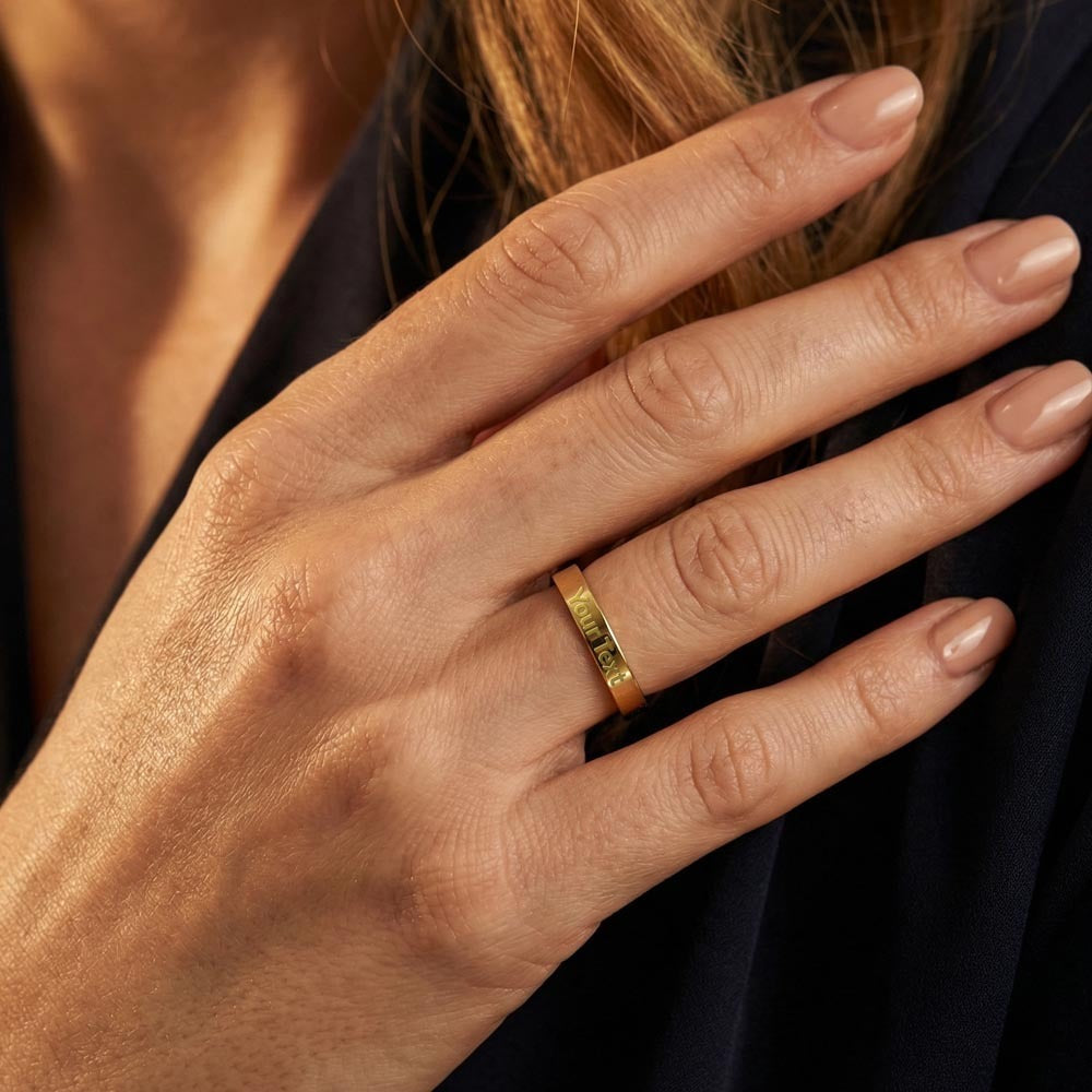 A close-up lifestyle photo of a woman's hand with a neutral-toned manicure resting elegantly over a black blouse, showcasing a polished gold flat-profile band ring on her ring finger. The custom ring features a clean, centered sample engraving that reads "Your Text" along the outer band, highlighting its personalized design.