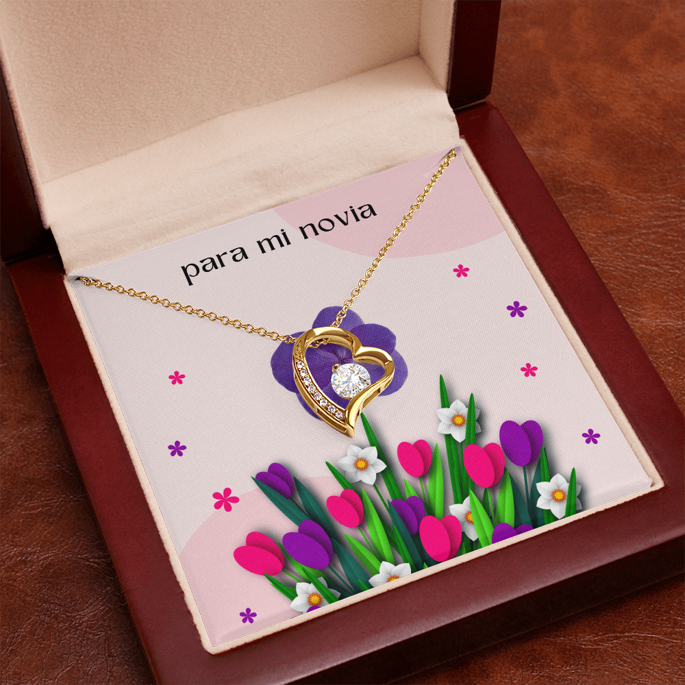 Collar de oro con dije de corazón y cristal brillante, presentado en una caja de madera de caoba abierta e inclinada con una tarjeta de regalo floral rosa que dice "para mi novia".