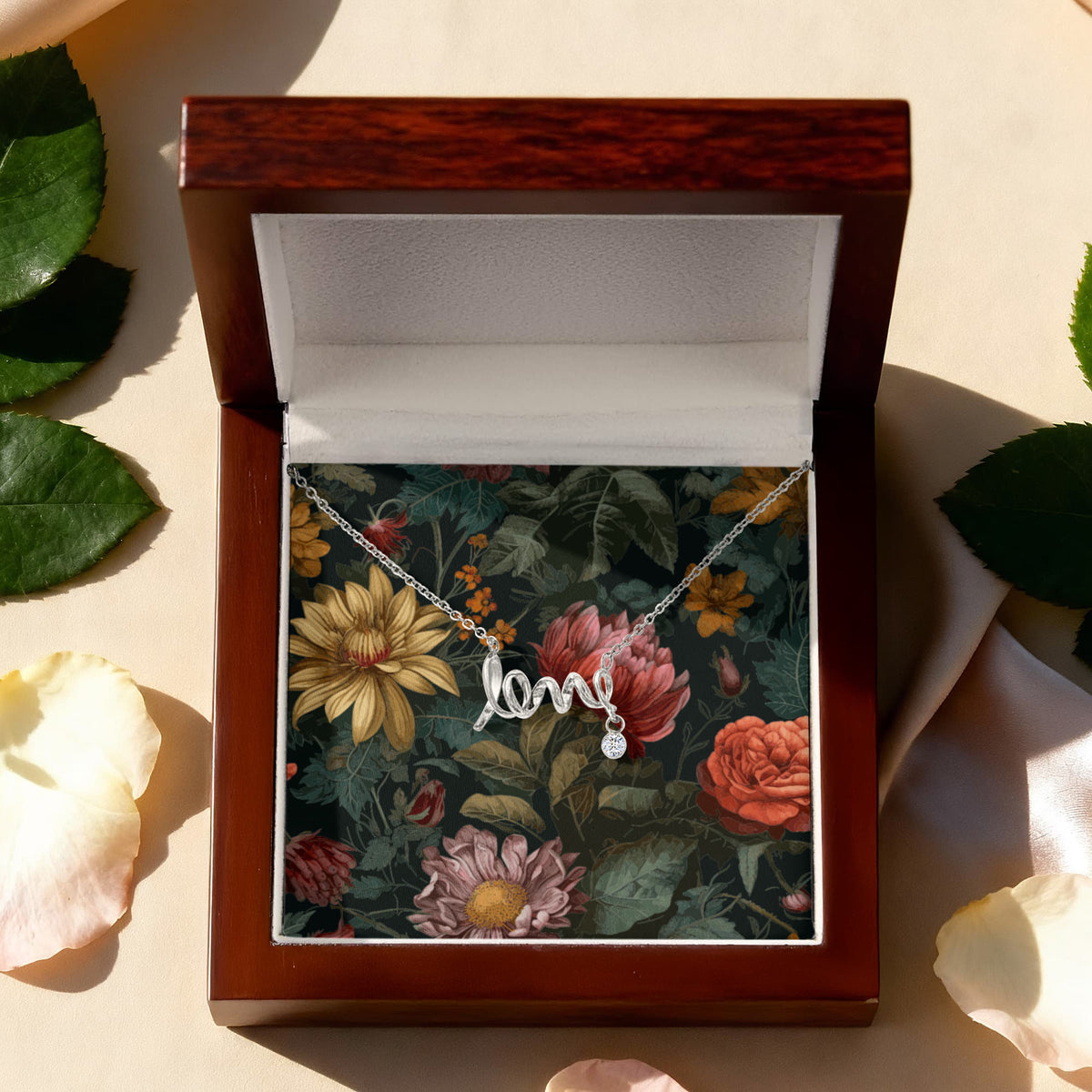 A top-down view of a silver script "love" necklace with a small dangling round crystal charm, displayed on a dark vintage floral card inside an open premium mahogany wooden gift box surrounded by scattered leaves and flower petals.