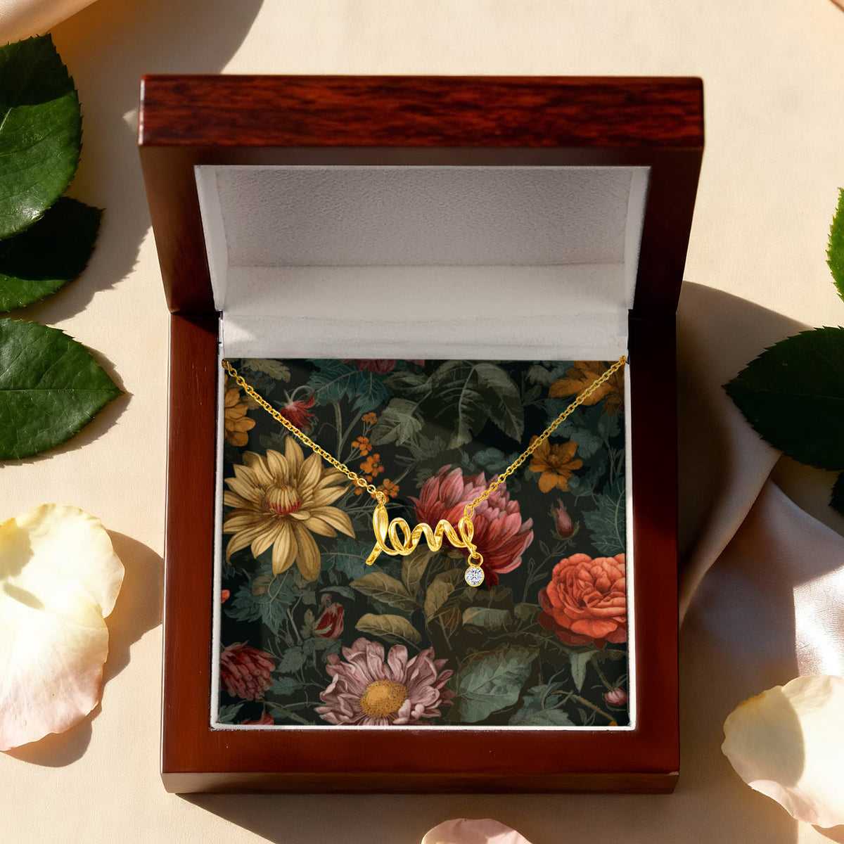 A top-down view of a gold script "love" necklace with a small dangling round crystal charm, displayed on a dark vintage floral card inside an open premium mahogany wooden gift box surrounded by scattered leaves and flower petals.