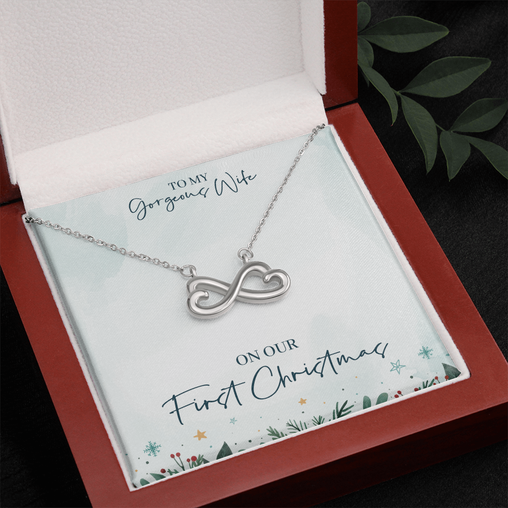 silver infinity heart necklace displayed in a dark red wood gift box. The presentation card features a festive holiday border with evergreens and stars, printed with the text: "TO MY Gorgeous Wife ON OUR First Christmas".