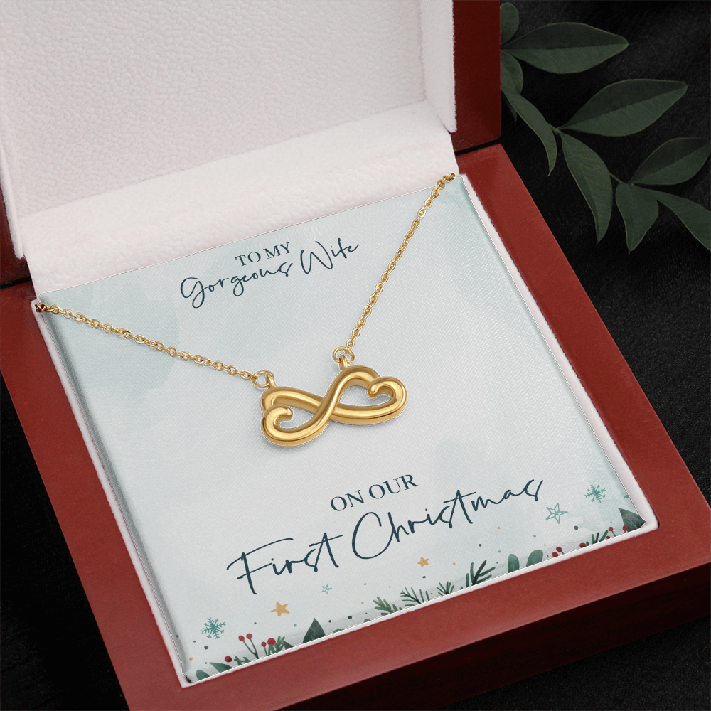 gold infinity heart necklace displayed in a dark red wood gift box. The presentation card features a festive holiday border with evergreens and stars, printed with the text: "TO MY Gorgeous Wife ON OUR First Christmas".
