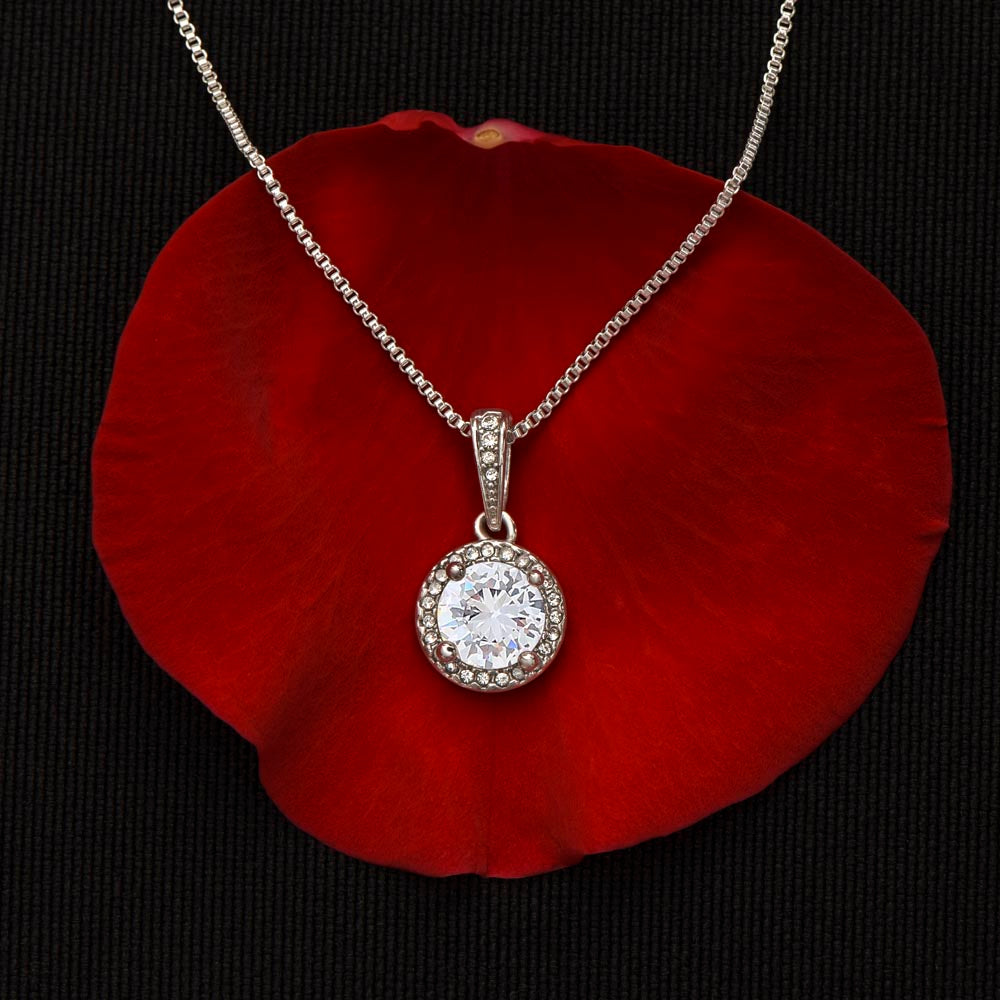 Close-up of a 14K white gold finish round cubic zirconia halo pendant necklace resting elegantly on a vibrant red rose petal against a dark black background to highlight the stone's sparkle and detail.
