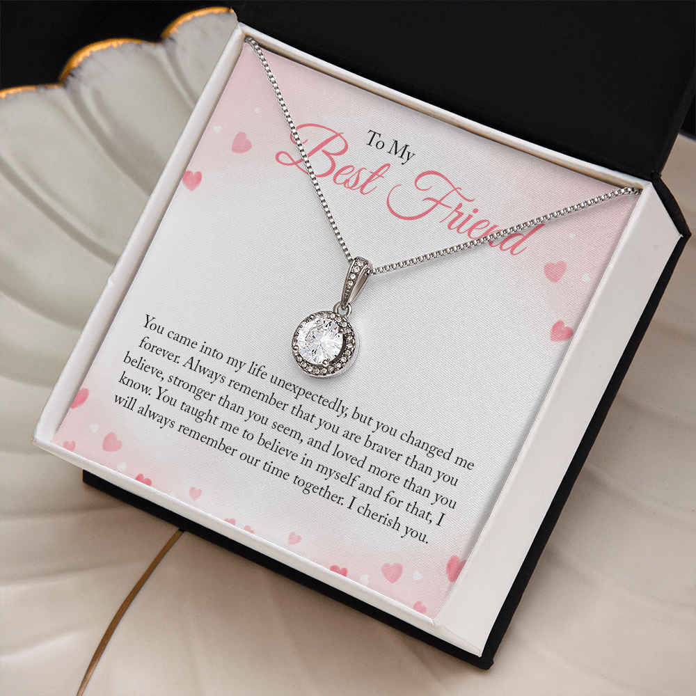 A round-cut cubic zirconia pendant necklace in white gold over a personalized 'To My Best Friend' sentimental message card with pink hearts, sitting on a cream colored shell shaped jewelry dish with gold trim.
