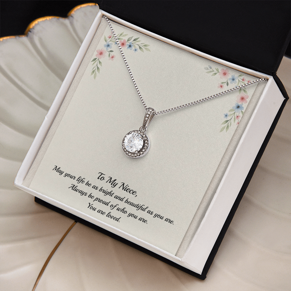 A silver necklace with a round bezel-set clear gemstone pendant, presented on a cream-colored gift card with floral corner details and a sentimental message for a niece, in a black and white two tone box sitting on a cream colored shell jewelry dish with gold trim.