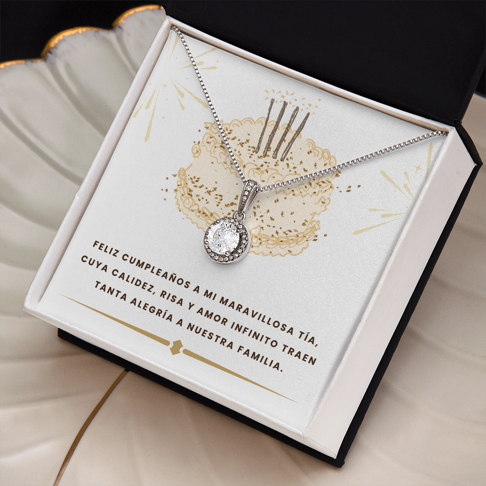 Vista en ángulo de un collar plateado con un dije circular de cristal brillante, colocado dentro de una caja de joyería blanca con bordes negros. El collar descansa sobre una tarjeta blanca decorada con un pastel dorado y el texto: "FELIZ CUMPLEAÑOS A MI MARAVILLOSA TÍA, CUYA CALIDEZ, RISA Y AMOR INFINITO TRAEN TANTA ALEGRÍA A NUESTRA FAMILIA".