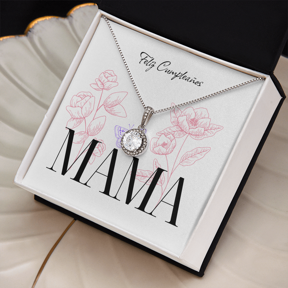 Vista en ángulo de un collar de plata con un dije redondo brillante, presentado dentro de una caja de regalo negra abierta. El collar descansa sobre una tarjeta blanca decorada con la frase "Feliz Cumpleaños" en letra cursiva, la palabra "MAMA" en letras grandes y negras, ilustraciones de flores rosadas y una pequeña mariposa morada.