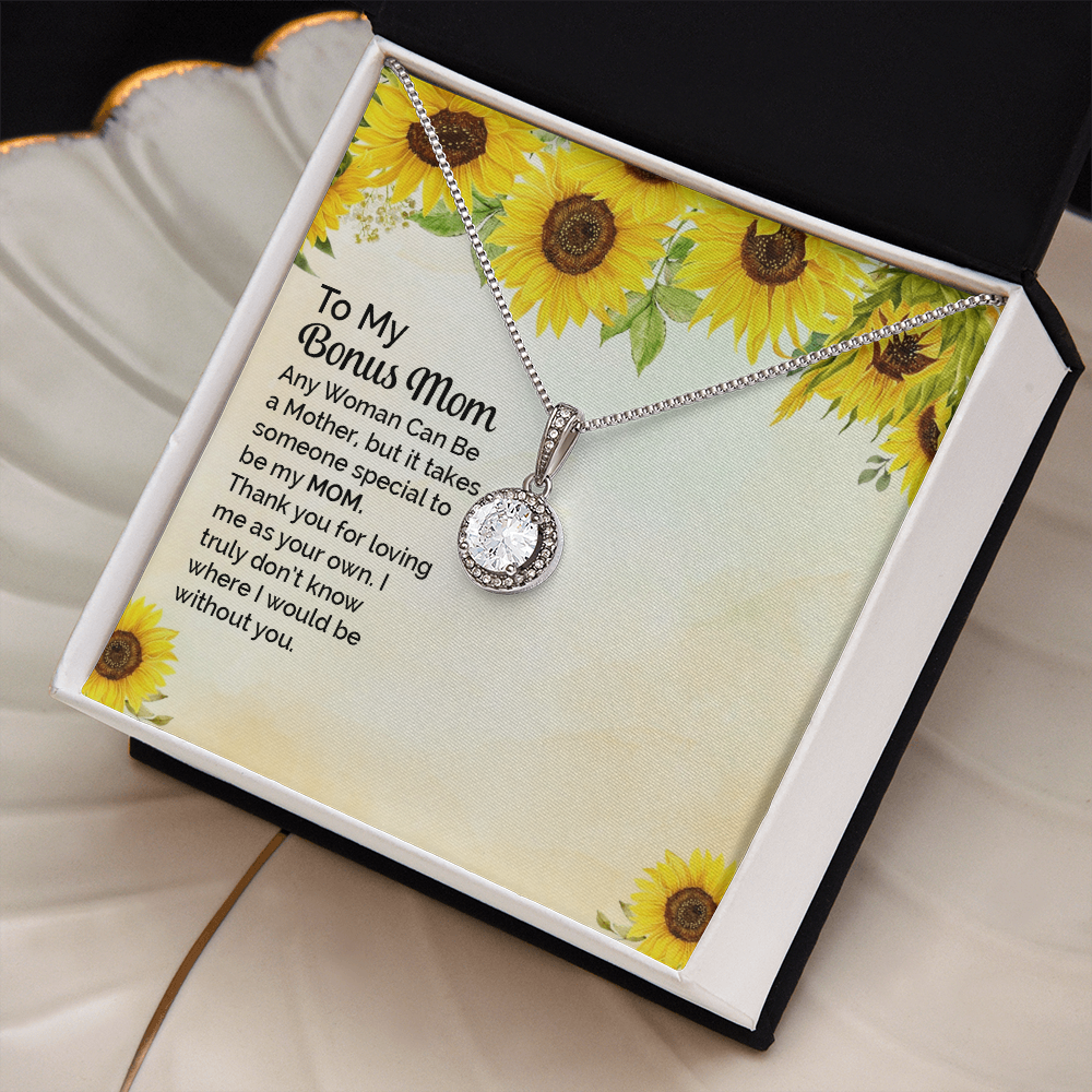 A silver necklace with a round bezel-set clear stone pendant is displayed on a printed gift card. The card has a sunflower border design and a sentimental message addressed to 'My Bonus Mom. The box sits on a cream colored shell jewelry plate with gold trim.
