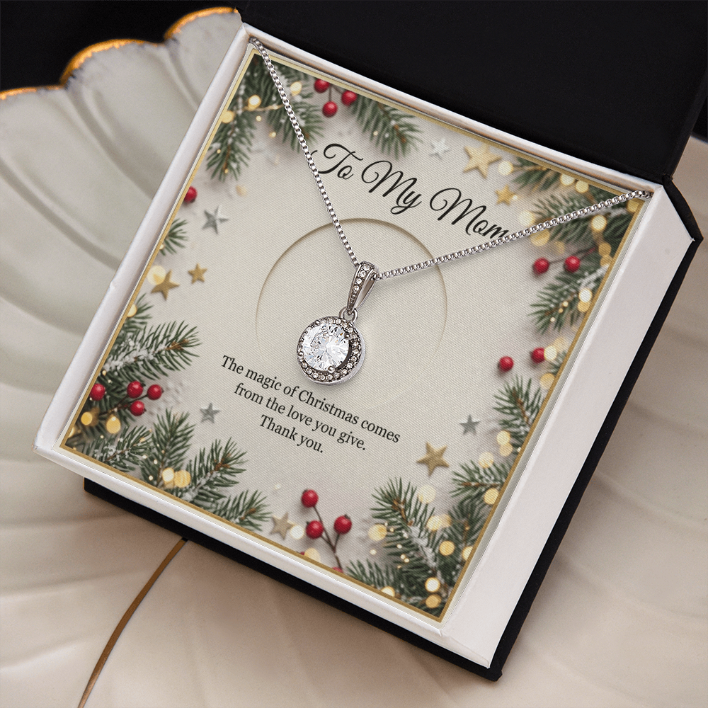 The card is framed by a festive border of evergreen pine branches, red winter berries, scattered gold and silver stars, and soft, glowing bokeh fairy lights. At the top of the insert card, elegant black cursive text reads "To My Mom." The silver necklace features a delicate chain that drapes downward to hold a round, brilliant-cut crystal pendant. The box lays on a cream colored, shell-shaped jewelry plate with gold trim.