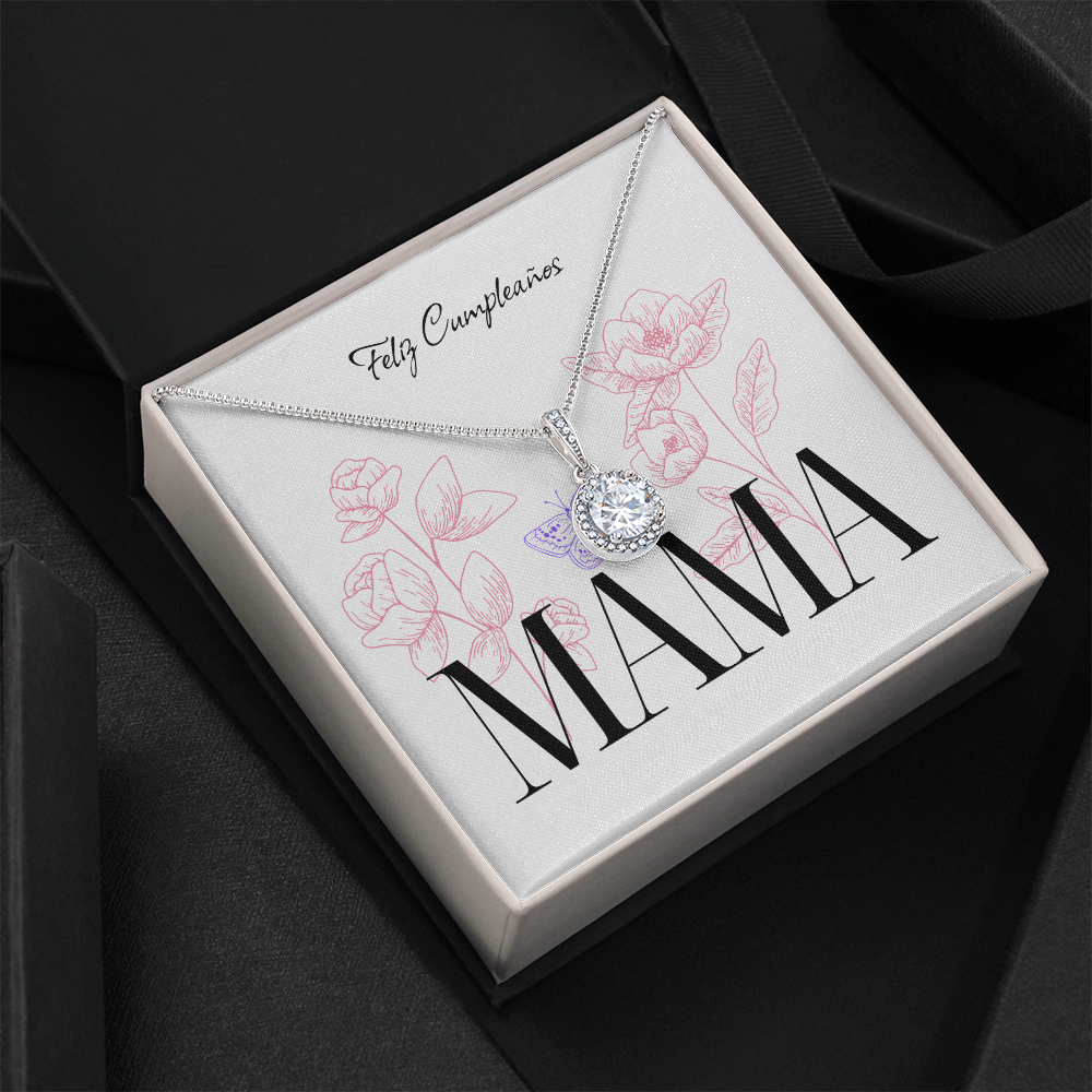 Vista elevada en ángulo de un collar de plata con dije redondo brillante dentro de su caja de regalo abierta, la cual está colocada sobre un fondo negro texturizado con una cinta de regalo oscura. La tarjeta blanca interior muestra el mensaje "Feliz Cumpleaños", la palabra "MAMA" en letras grandes, diseños de flores rosadas y una mariposa morada.
