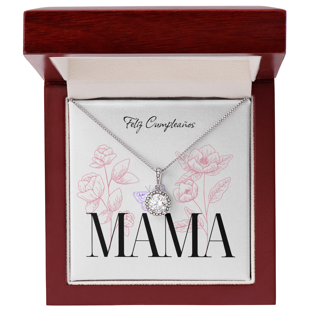 Collar plateado con un brillante dije circular de cristal rodeado de pequeñas gemas, presentado dentro de un estuche de madera color caoba abierto. El collar está montado sobre una tarjeta blanca decorada con delicadas ilustraciones de flores rosas y una pequeña mariposa morada. En la parte superior de la tarjeta se lee "Feliz Cumpleaños" en letra cursiva, y en la parte inferior resalta la palabra "MAMÁ" en letras grandes y oscuras.
