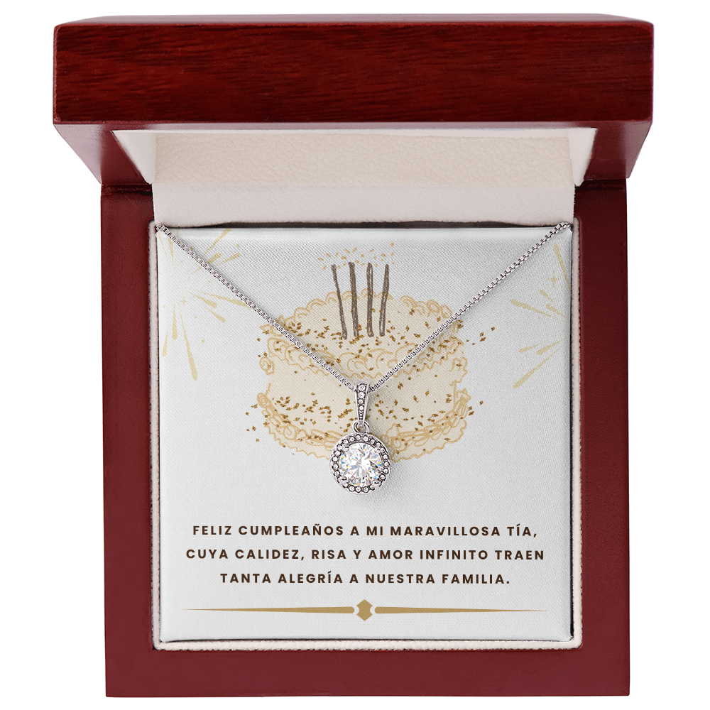 Collar plateado con un brillante dije circular de cristal, colocado de frente dentro de un elegante estuche de madera color caoba abierto. El collar está montado sobre una tarjeta blanca ilustrada con un pastel dorado y la dedicatoria en letras oscuras: "FELIZ CUMPLEAÑOS A MI MARAVILLOSA TÍA, CUYA CALIDEZ, RISA Y AMOR INFINITO TRAEN TANTA ALEGRÍA A NUESTRA FAMILIA".
