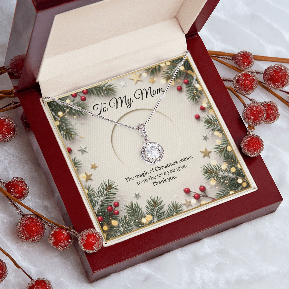 The card is framed by a festive border of evergreen pine branches, red winter berries, scattered gold and silver stars, and soft, glowing bokeh fairy lights. At the top of the insert card, elegant black cursive text reads "To My Mom." The silver necklace features a delicate chain that drapes downward to hold a round, brilliant-cut crystal It sits in a dark red wood gift box that sits on a white table surrounded by red Christmas berries.