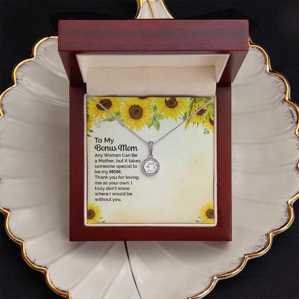 A silver necklace with a round bezel-set clear stone pendant is displayed on a printed gift card. The card has a sunflower border design and a sentimental message addressed to 'My Bonus Mom. The piece sits in a dark red wood gift box that sits on a cream colored shell-shaped jewelry dish with gold trim.