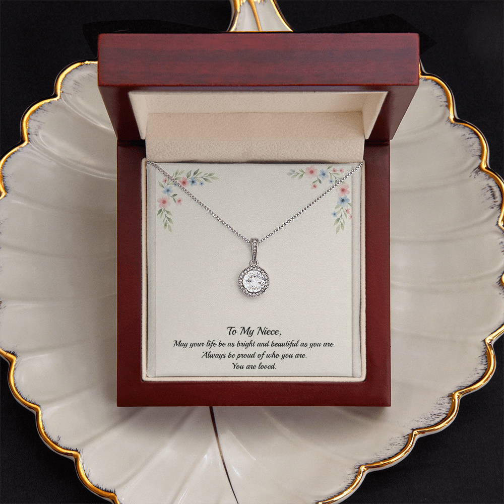 A silver necklace with a round bezel-set clear gemstone pendant, presented on a cream-colored gift card with floral corner details and a sentimental message for a niece, in a dark red wood box sitting on a cream colored shell jewelry dish with gold trim.