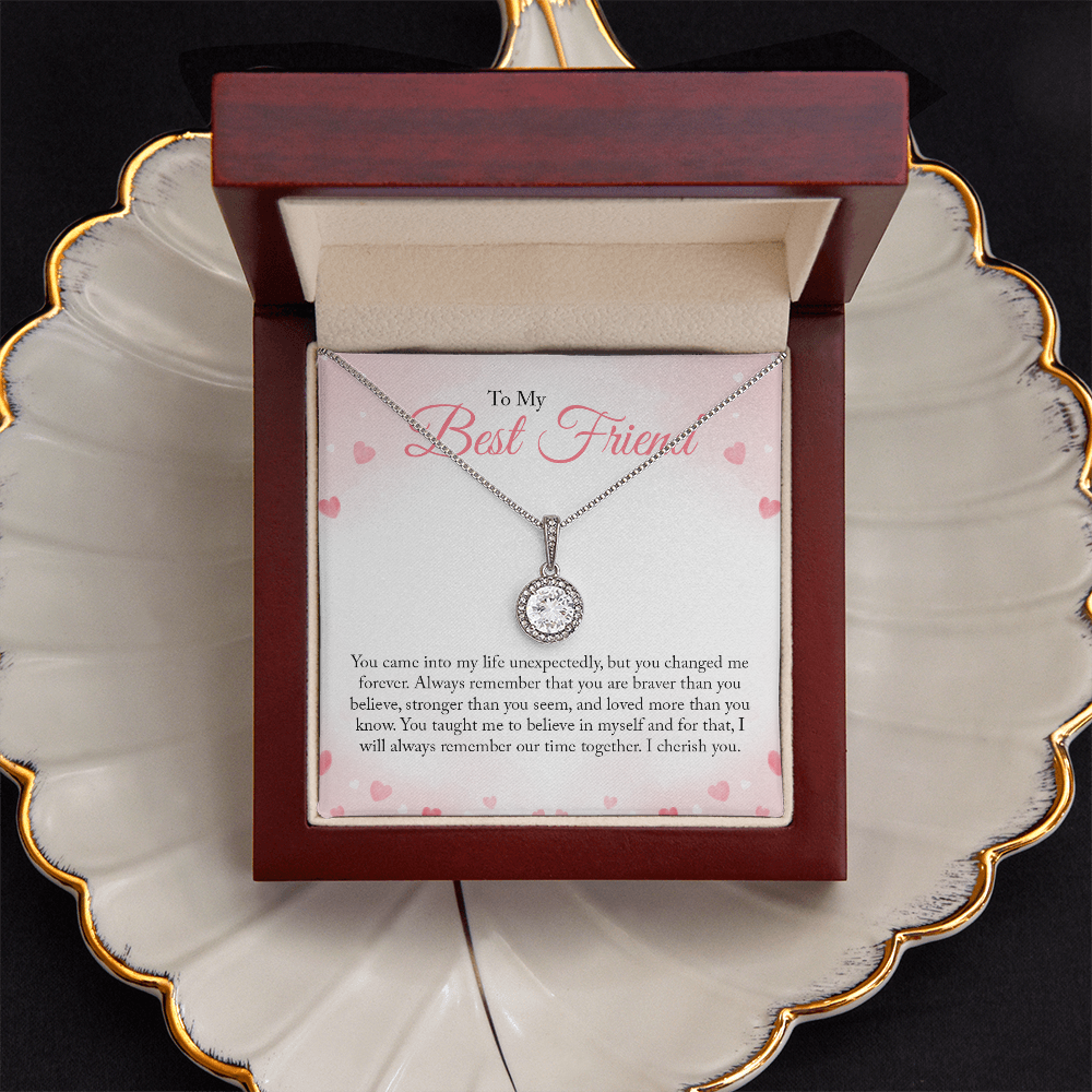 A round-cut cubic zirconia pendant necklace in white gold over a personalized 'To My Best Friend' sentimental message card with pink hearts in a dark red wood gift box that sits on a cream colored shell shaped jewelry dish with gold trim.