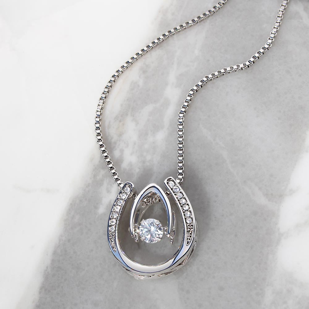 Close up of a silver lucky horseshoe necklace with cubic zirconia laying on a gray and white marble surface.