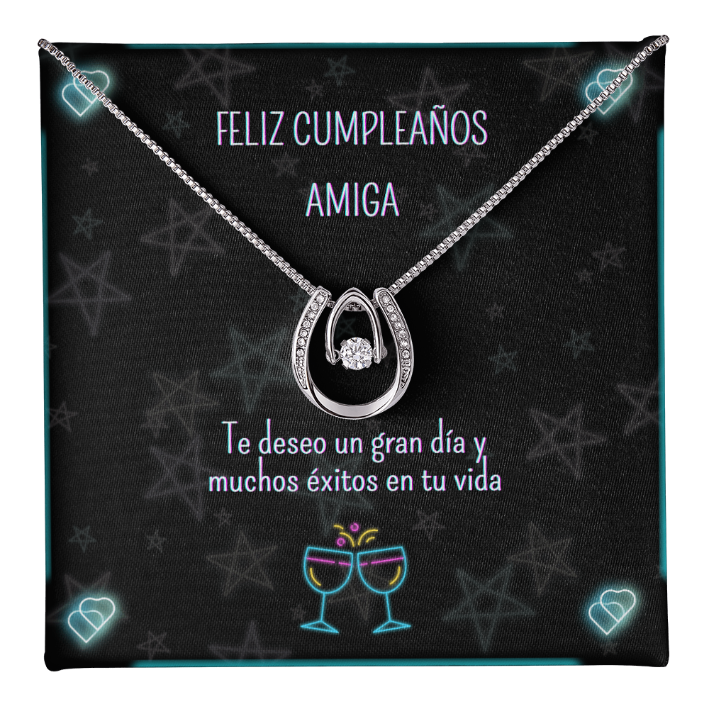 Collar plateado con un colgante en forma de herradura y una piedra brillante central, colocado sobre una tarjeta de regalo negra decorada con estrellas y luces de neón que lleva el mensaje en español: "Feliz Cumpleaños Amiga. Te deseo un gran día y muchos éxitos en tu vida".