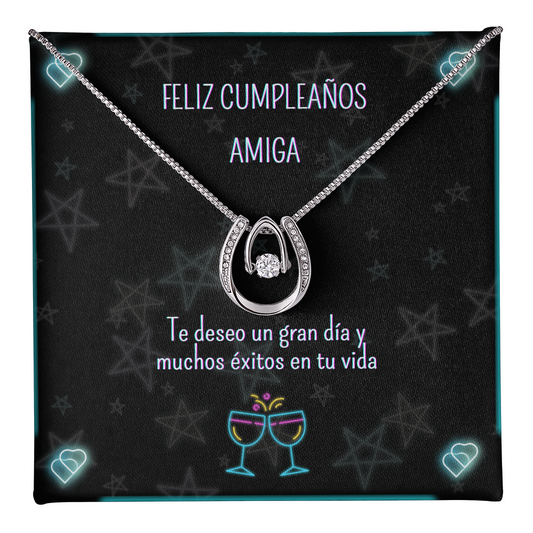 Collar plateado con un colgante en forma de herradura y una piedra brillante central, colocado sobre una tarjeta de regalo negra decorada con estrellas y luces de neón que lleva el mensaje en español: "Feliz Cumpleaños Amiga. Te deseo un gran día y muchos éxitos en tu vida".