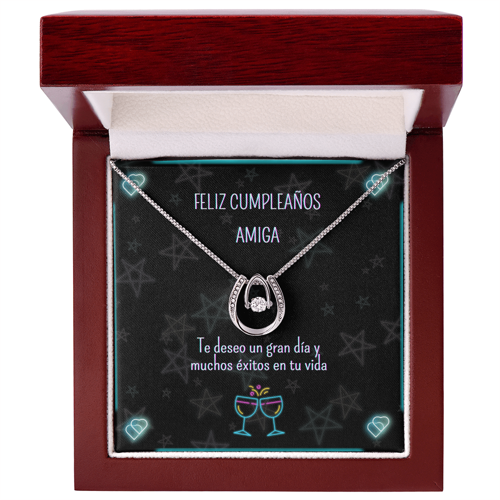 Collar de herradura plateado con piedra brillante central, presentado dentro de una caja de regalo de madera de lujo abierta con acabado de caoba. La tarjeta interior muestra estrellas de fondo y el mensaje en español: "Feliz Cumpleaños Amiga. Te deseo un gran día y muchos éxitos en tu vida".