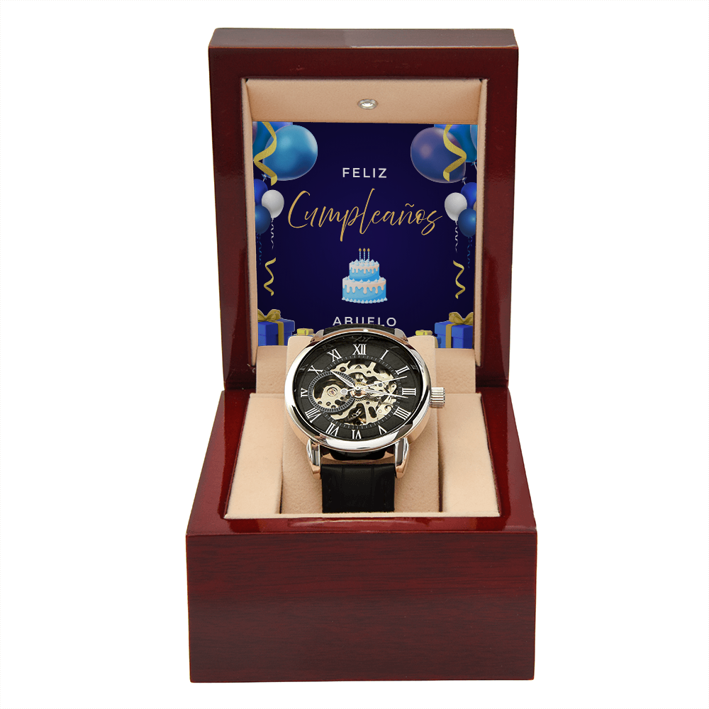 Reloj esqueleto de lujo para hombre con correa negra, presentado dentro de una caja de madera de caoba abierta con una tarjeta de feliz cumpleaños para abuelo de fondo.