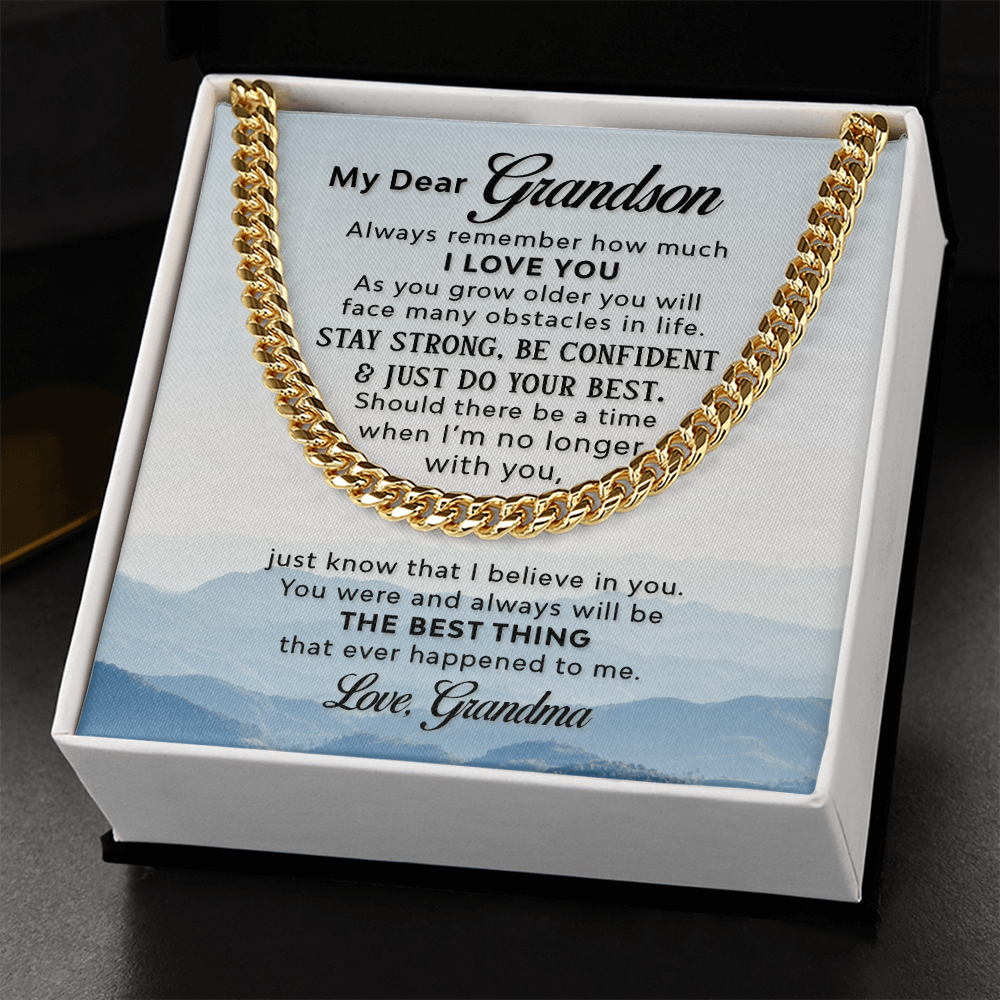 A gold-colored men's Cuban link chain necklace draped over a square white message card inside a wooden gift box. The background of the card features a scenic, misty blue mountain landscape. The gift box sits on a black.