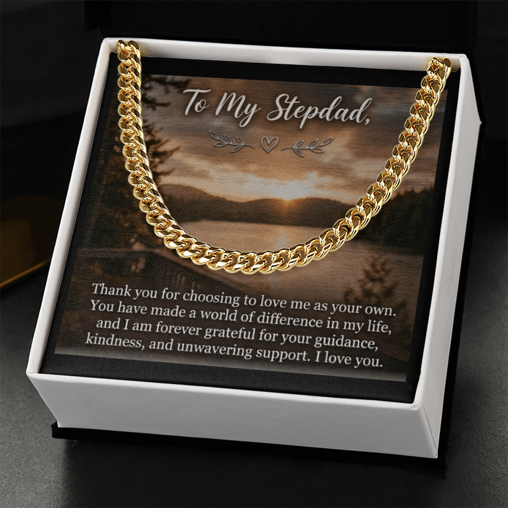 The card displays a scenic lakeside landscape at sunset or sunrise with dense pine trees lining the shores under a cloudy sky. At the top, the elegant script text reads, "To My Stepdad," adorned with a small heart and leaf flourishes underneath. The bottom half of the card features a heartfelt paragraph printed in a clean, white serif font. The box rests on a black surface and the gift box features a gold men's Cuban link chain necklace.