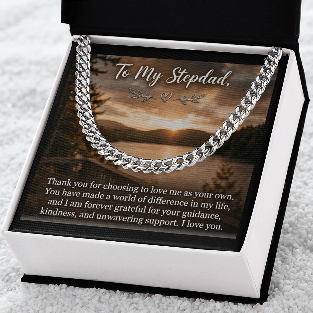 The black gift box features a plush white interior frame that highlights the square insert card. The card displays a scenic lakeside sunset landscape with a forested shoreline. At the top of the card, text in an elegant cursive font reads, "To My Stepdad," accented by small heart and leaf illustrations directly below it. Printed clearly across the lower section of the card is a heartfelt message in white serif text. A silver-toned men's cuban link chain rests on the image.