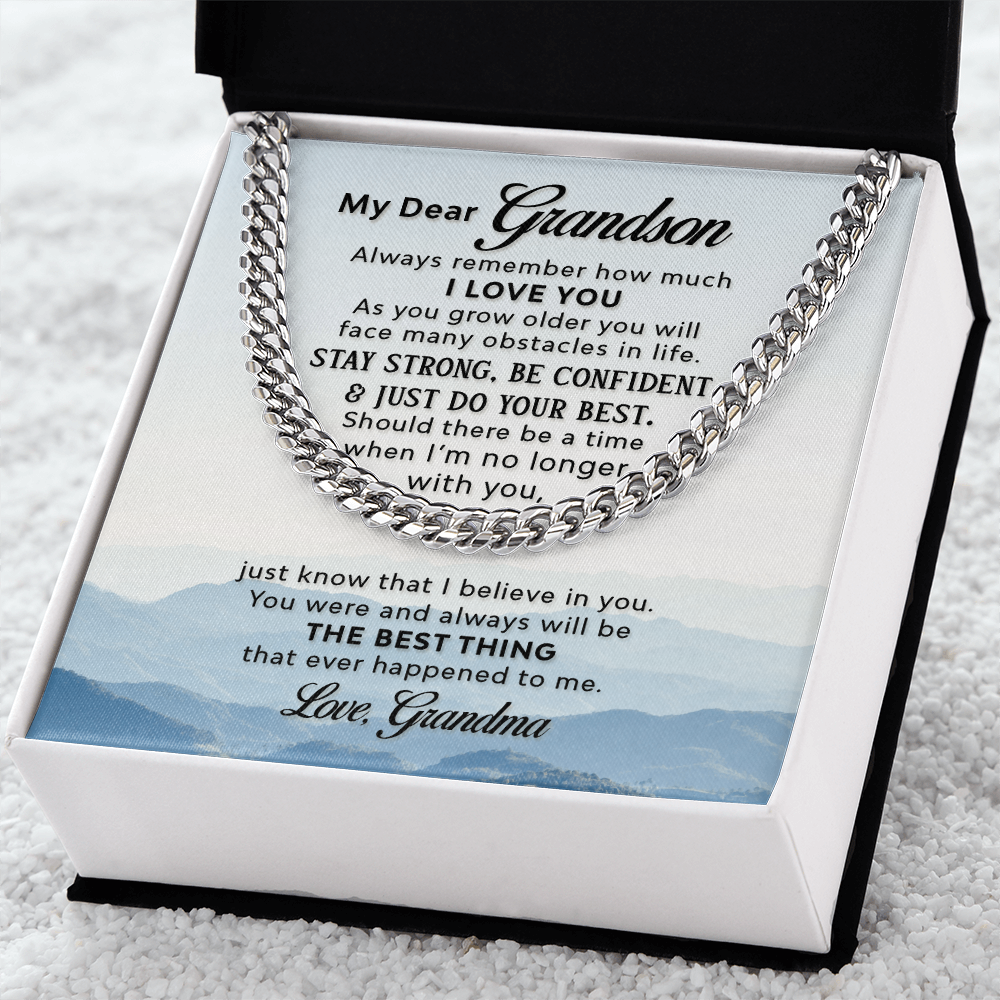 A silver-colored men's Cuban link chain necklace draped over a square white message card inside a gift box. The background of the card features a scenic, misty blue mountain landscape, and the gift box lays on a white towel like fabric.