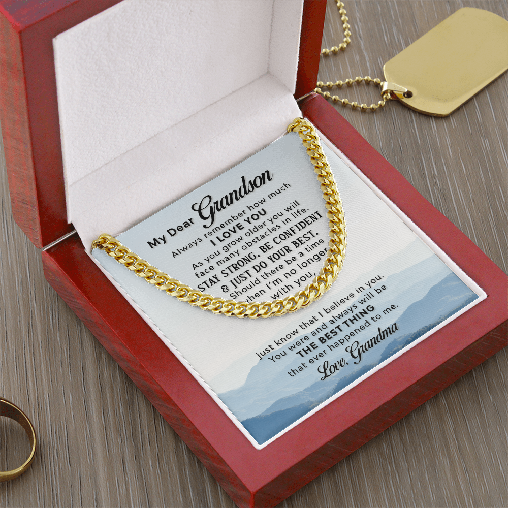 A gold-colored men's Cuban link chain necklace draped over a square white message card inside a wooden gift box. The background of the card features a scenic, misty blue mountain landscape. The gift box sits on a light gray table surrounded with a gold ring and gold dog tag necklace.