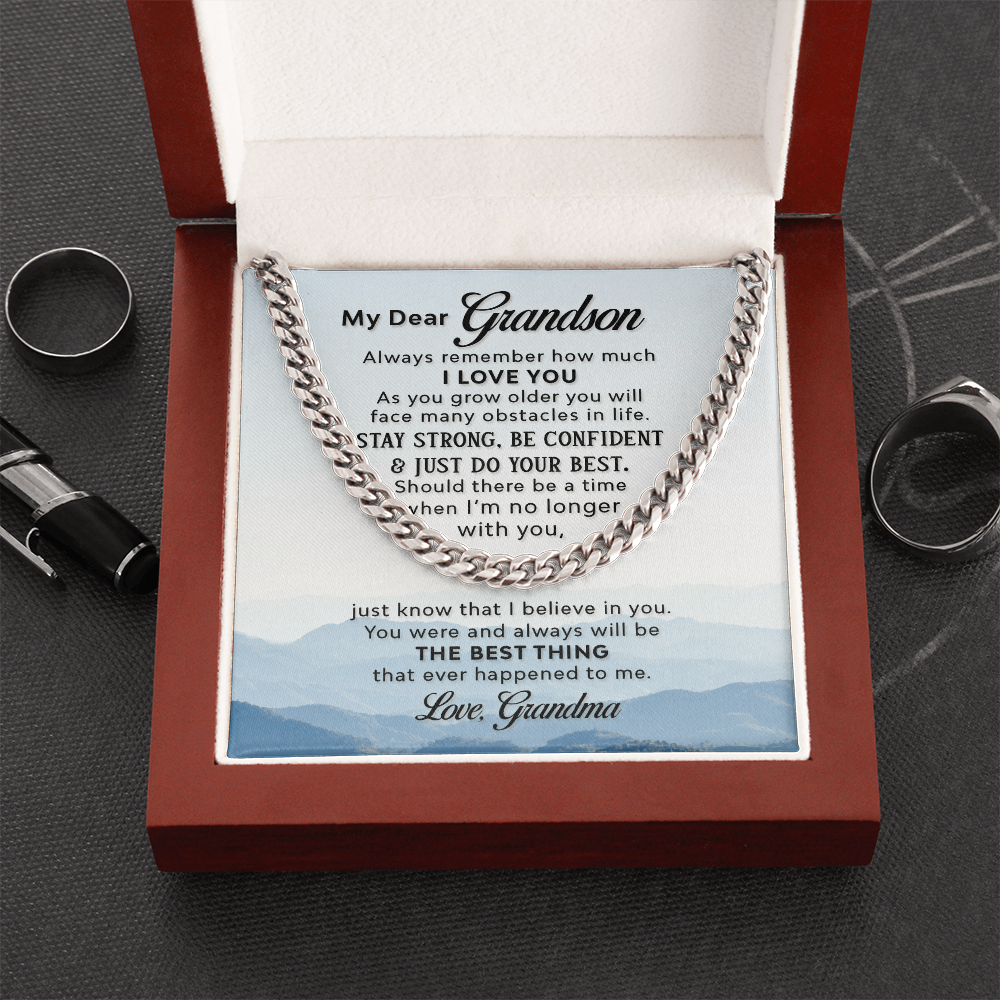 A silver-colored men's Cuban link chain necklace draped over a square white message card inside a wooden gift box. The background of the card features a scenic, misty blue mountain landscape. The gift box sits on a dark gray table surrounded with a pen and 2 silver men's rings.